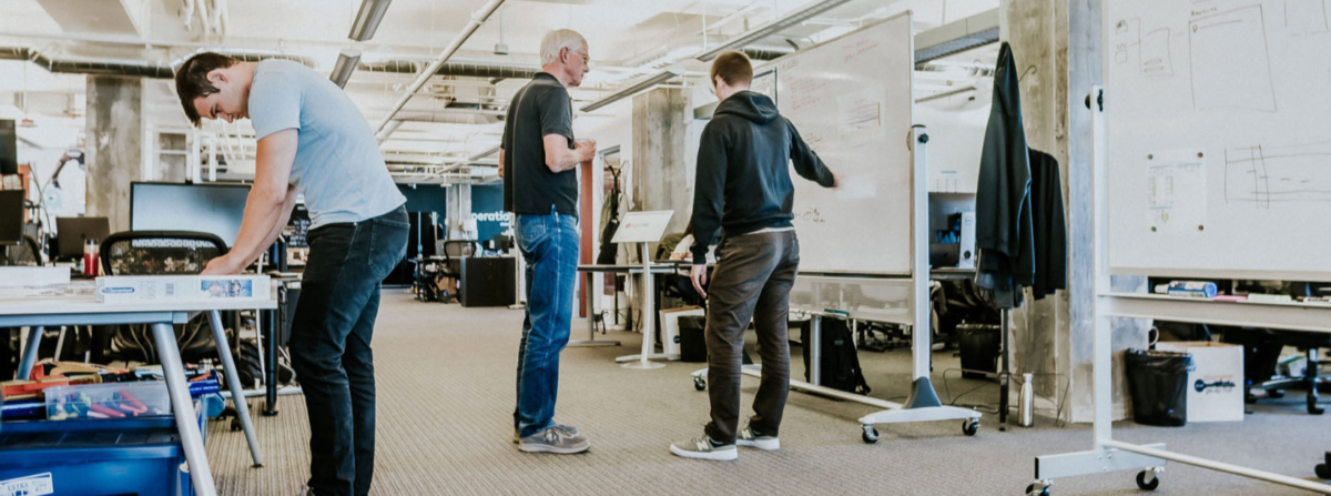 3 men in an office, one building at a table, two talking at a whiteboard.